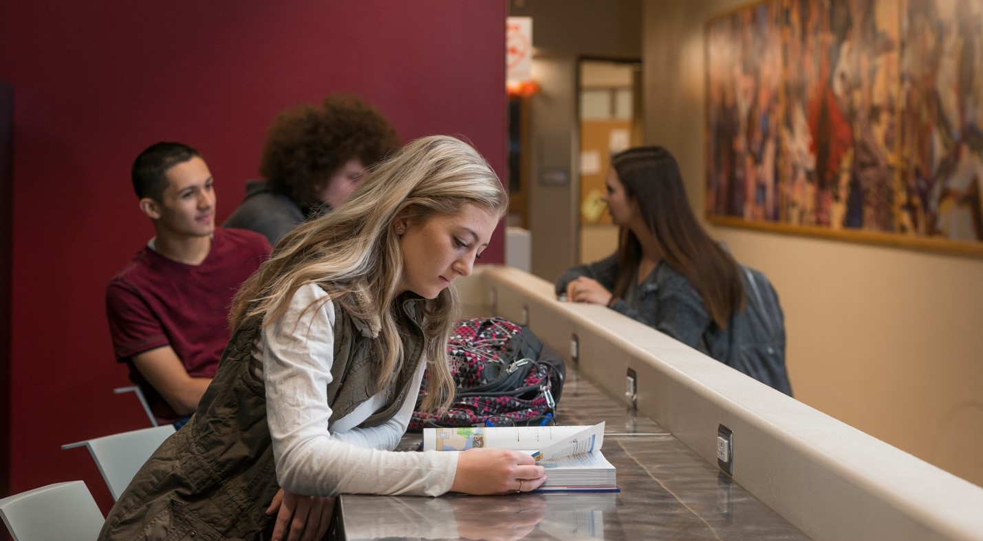 Several PCC students sitting at a counter against a maroon back wall, one woman standing opposite partially in front of a mural just visible in the background; the young woman in the foreground is reading a book at the counter.